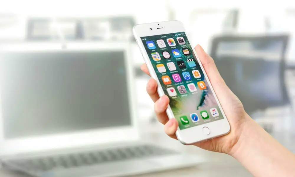 Close-up view of a smartphone displaying apps, held by a hand, with a blurred laptop in the background.